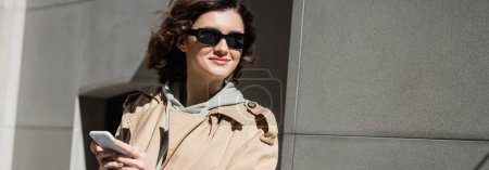 begeisterte und stilvolle Frau mit dunkler Sonnenbrille, beigem Trenchcoat und Kapuzenpullover, während sie in der Nähe eines grauen Gebäudes auf der städtischen Straße der europäischen Stadt steht, Banner