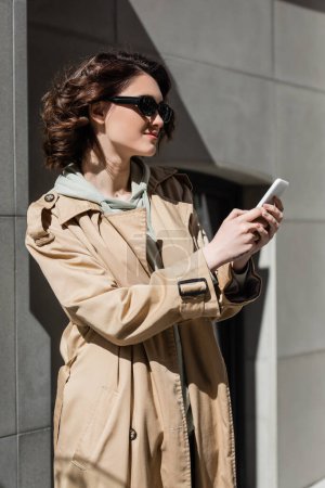 femme joyeuse avec des lunettes de soleil sombres et élégantes et un trench coat beige, avec des cheveux bruns ondulés et un tatouage bavardant sur smartphone près du bâtiment gris en plein soleil dans la rue urbaine, mode de vie de voyage