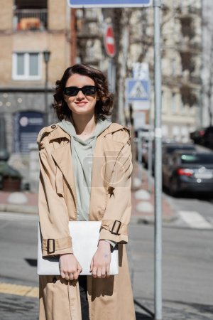 carefree woman with wavy brunette hair and tattoo, wearing beige trench coat, grey hoodie and dark sunglasses while standing with laptop on urban street of european city on blurred background