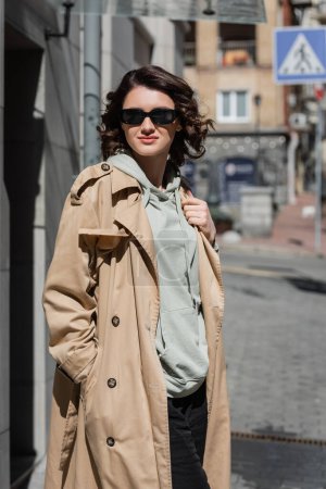 mujer encantadora y de moda en gafas de sol oscuras, sudadera con capucha gris y gabardina beige de pie en la ciudad europea sobre un fondo borroso, fotografía callejera, estilo de vida de viaje 