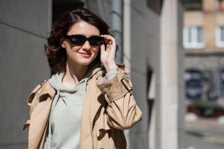 Photo for Young and pleased woman with wavy brunette hair, in grey hoodie and beige trench coat adjusting dark stylish sunglasses and looking away on urban street on blurred background - Royalty Free Image