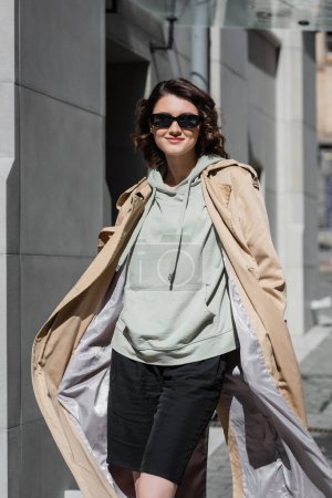 mujer elegante y despreocupada en gafas de sol oscuras, sudadera con capucha gris y gabardina beige posando en la calle en la ciudad europea moderna y mirando a la cámara, estilo de vida de viaje, moda urbana