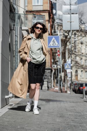 full length of young confident woman in dark stylish sunglasses, grey hoodie, beige trench coat and black shorts holding hand in pocket and walking in European city, street photography, travel 