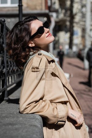 Photo for Side view of young and carefree woman with wavy brunette hair and tattoo, in dark sunglasses and beige trench coat relaxing near forged fence in european city, street photography, urban fashion - Royalty Free Image