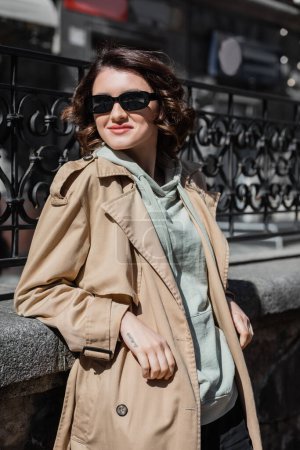 charismatic tattooed woman with wavy brunette hair, in dark stylish sunglasses, grey hoodie and beige trench coat smiling and looking away near forged fence in city, street photography