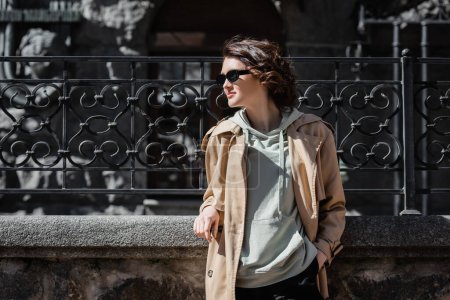 travel lifestyle, young and fashionable woman with wavy brunette hair, in dark sunglasses, beige trench coat and grey hoodie looking away while standing near forged fence on city street, travel lifestyle