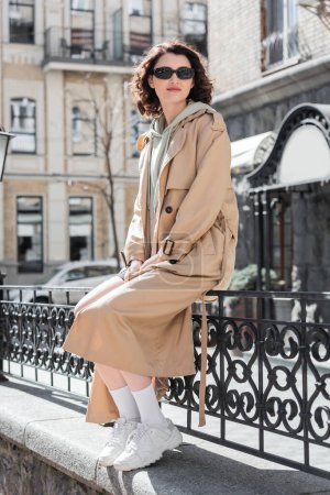 full length of trendy young woman in stylish casual clothes, beige trench coat, white sneakers and dark sunglasses sitting on forged fence and looking away on street of European city