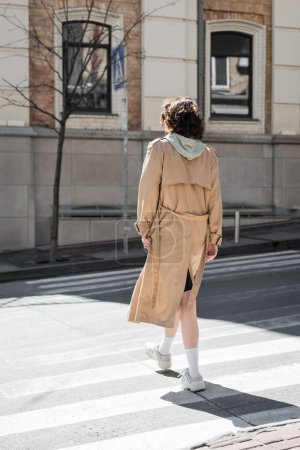 full length of young woman with wavy brunette hair, in beige trench coat, grey hoodie and white sneakers crossing road on sunny street of European city, back view, urban fashion, travel lifestyle 