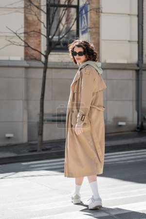 Photo for Full length of young and stylish woman with wavy brunette hair crossing road in dark sunglasses, grey hoodie, beige trench coat and white sneakers and looking at camera in European city - Royalty Free Image