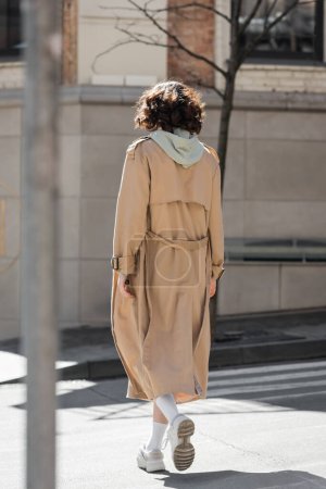 back view of young woman with wavy brunette hair, in stylish casual clothes, grey hoodie, beige trench coat and white sneakers  crossing road on street of modern European city, urban lifestyle