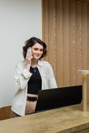 Photo for Happy young receptionist with wavy brunette hair and tattoo, in stylish casual clothes talking on mobile phone while working at front desk near computer monitor and lamp in hotel lobby - Royalty Free Image
