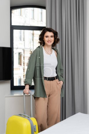 Photo for Smiling woman with wavy brunette hair and in stylish clothes looking at camera while standing with hand in pocket near window, grey curtains, lcd tv and bed in cozy hotel suite - Royalty Free Image