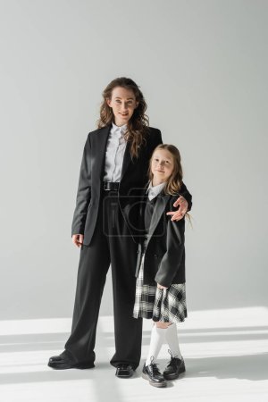 moderne Mutter und Tochter, Geschäftsfrau im Anzug umarmt Schulmädchen in Uniform mit kariertem Rock, auf grauem Hintergrund, Blazer, sich auf das neue Schuljahr vorbereiten, in die Kamera schauen, formelle Kleidung 