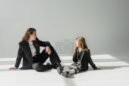Photo for Fashionable mother and daughter, businesswoman in suit sitting and looking at schoolgirl in uniform with plaid skirt on grey background, blazers, new school year, looking at each other - Royalty Free Image