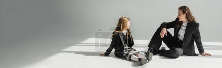 Photo for Modern mother and child, businesswoman in suit sitting and looking at schoolgirl in uniform with plaid skirt on grey background, blazers, new school year, looking at each other, banner - Royalty Free Image