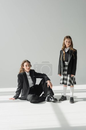 working mother and child, businesswoman in suit sitting near schoolgirl in uniform with plaid skirt on grey background, blazers, new school year, looking at camera, formal attire 