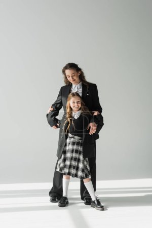 Foto de Crianza moderna, mujer de negocios de moda en traje abrazando a la hija en uniforme escolar con falda a cuadros y de pie juntos sobre fondo gris, madre feliz y el niño - Imagen libre de derechos