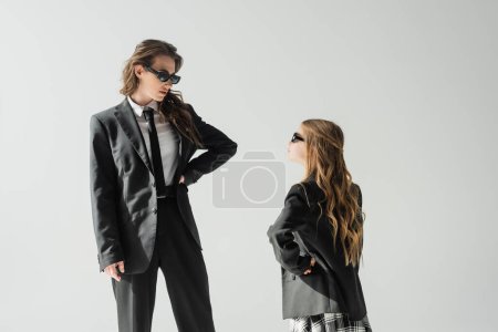 Foto de Madre e hija con estilo en gafas de sol, mujer de negocios en traje y colegiala en uniforme mirándose entre sí mientras están de pie sobre fondo gris en el estudio, la familia de moda - Imagen libre de derechos