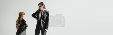 Photo for Modern mother and daughter in sunglasses, businesswoman in suit and schoolgirl in uniform looking at each other while standing on grey background in studio, fashionable family, banner - Royalty Free Image