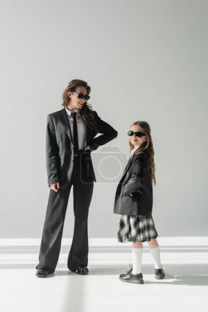 mujer alegre con su hija, mujer de negocios en traje posando con la mano en la cadera y colegiala en gafas de sol y uniforme de pie juntos sobre fondo gris en el estudio, atuendo formal 
