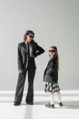 cheerful woman with her daughter, businesswoman in suit posing with hand on hip and schoolgirl in sunglasses and uniform standing together on grey background in studio, formal attire  puzzle #659557810