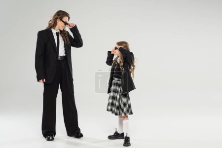 stilvolle Mutter und Kind mit Sonnenbrille, glückliche Geschäftsfrau in formeller Kleidung, die Schulmädchen in Uniform auf grauem Hintergrund im Atelier anschaut, modische Familie, moderne Elternschaft 