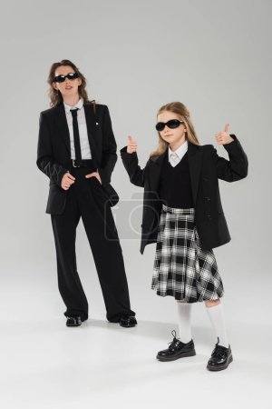stylish schoolgirl in sunglasses and uniform showing thumbs up and standing near modern mother on grey blurred background in studio, formal attire, back to school 