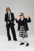 stylish schoolgirl in sunglasses and uniform showing thumbs up and standing near modern mother on grey blurred background in studio, formal attire, back to school  Poster #659557866