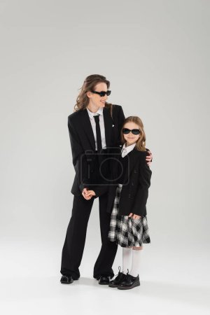 Moderne Elternschaft, fröhliche Geschäftsfrau in Anzug und Sonnenbrille umarmt Tochter in Schuluniform mit kariertem Rock und steht zusammen auf grauem Hintergrund, Mutter und Kind 