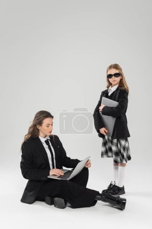 digitaler Nomadentum, modische Frau im Anzug mit Laptop in der Nähe der Tochter in Schuluniform und Sonnenbrille auf grauem Hintergrund, Fernarbeit, berufstätige Mutter, Geschäftskleidung 