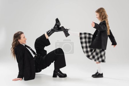 Photo for Modern parenting, having fun, side view of happy mother in suit and child in school uniform touching boots of each other on grey background in studio, blazer, businesswoman, back to school - Royalty Free Image