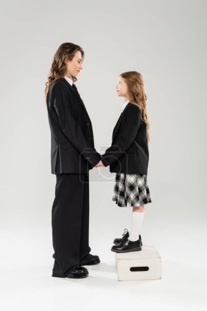 mother and child holding hands, happy woman in business attire and child in school uniform standing on step stool on grey background, modern parenting, face to face