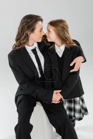 happy mother hugging daughter, businesswoman sitting on concrete stool near kid in uniform on grey background in studio, formal attire, modern parenting, fashion shoot 