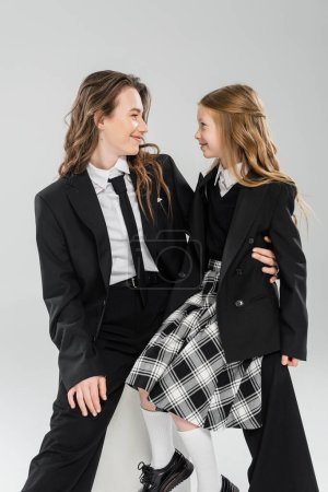 Photo for Working mother and daughter, happy businesswoman sitting on concrete stool near kid in uniform on grey background in studio, formal attire, modern parenting, fashion shoot - Royalty Free Image