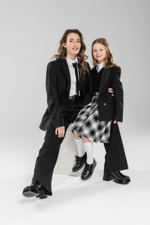 Photo for Working mother and daughter, happy businesswoman and schoolgirl sitting on concrete stool together on grey background in studio, formal attire, modern parenting, fashion shoot - Royalty Free Image