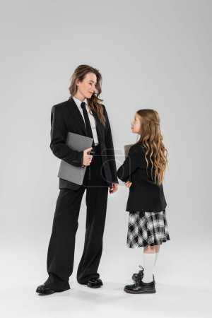 working mother and daughter, digital nomadism, remote work, e learning, businesswoman in suit and schoolgirl standing together with laptops on grey background in studio, modern parenting 