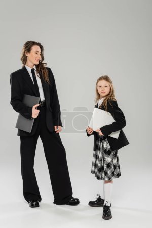 Mutter und Tochter, digitaler Nomadentum, Fernarbeit, E-Learning, glückliche Geschäftsfrau im Anzug und Schulmädchen, die mit Laptops auf grauem Hintergrund im Studio stehen, moderne Elternschaft 