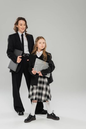 happy mother and daughter, digital nomadism, remote work, e learning, businesswoman in suit and schoolgirl standing together with laptops on grey background in studio, modern parenting 