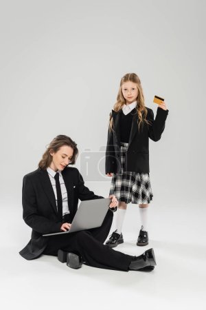 working mother and daughter, digital nomadism, schoolgirl holding credit card near mom using laptop on grey background, modern parenting, business attire, financial education, online purchase