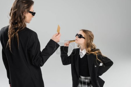 mother and daughter in sunglasses, businesswoman in suit and schoolgirl in uniform holding credit cards and looking at each other on grey background, modern parenting, money management 