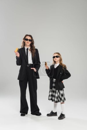 mother and daughter in sunglasses, businesswoman in suit and schoolgirl in uniform holding credit cards on grey background, modern parenting, financial learning, budgeting, money management  