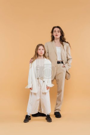 stylish mother and daughter in suits, woman and girl looking at camera while standing together on beige background, fashionable outfits, formal attire, corporate mom, modern family, hand in pocket  