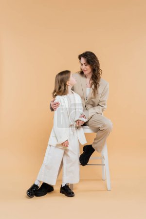 stylish mother and daughter in suits, woman sitting on high chair and looking at girl on beige background, fashionable outfits, formal attire, corporate mom, modern family 