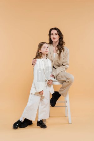 stylish mother and daughter in suits, woman and girl looking at camera, sitting on high chair on beige background, fashionable outfits, formal attire, corporate mom, modern family 