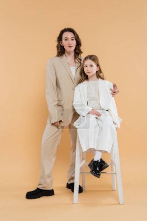 madre e hija con estilo en trajes, mirando a la cámara mientras está sentado en la silla alta cerca de la mujer en fondo beige, trajes de moda, atuendo formal, mamá corporativa, familia moderna 