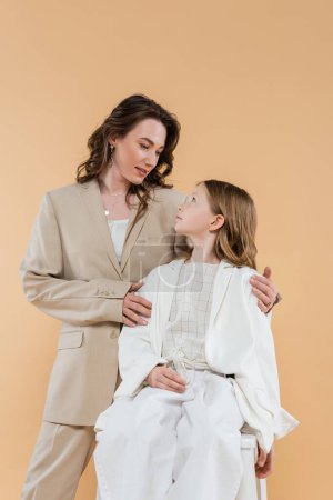 madre e hija de negocios en trajes, mujer abrazando hombros de niña sentada en silla sobre fondo beige, trajes de moda, atuendo formal, mamá corporativa, familia moderna 