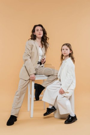 Photo for Corporate mom and daughter in suits, woman sitting on high chair and looking at camera near girl on beige background, fashionable outfits, formal attire, corporate mom, motherhood and career - Royalty Free Image