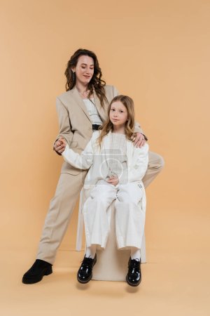 Photo for Stylish mother and daughter in suits, woman and girl holding hands and sitting together on beige background, fashionable outfits, formal attire, corporate mom, modern family - Royalty Free Image