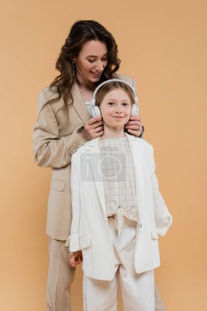 Photo for Trendy mother and daughter in suits, woman wearing wireless headphones on girl while standing together on beige background, fashionable outfits, formal attire, corporate mom, modern family - Royalty Free Image