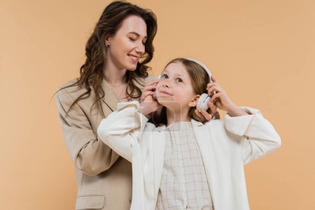 Photo for Corporate mom and daughter in suits, happy woman wearing wireless headphones on girl while standing together on beige background, fashionable outfits, formal attire, modern family - Royalty Free Image